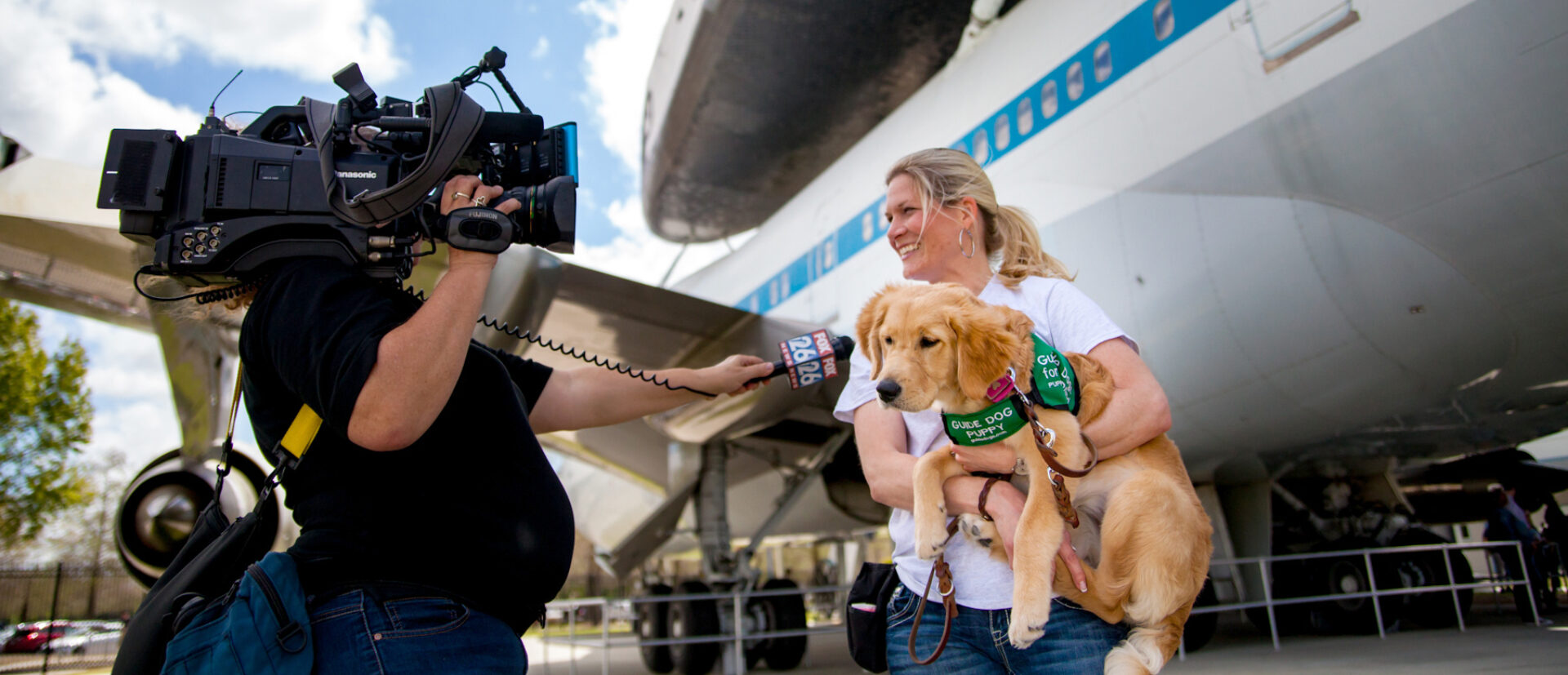 A television reporter interviews a Guide Dogs for the Blind puppy raising volunteer who holds a Golden Retriever guide dog puppy in front of the space shuttle at the NASA Museum.