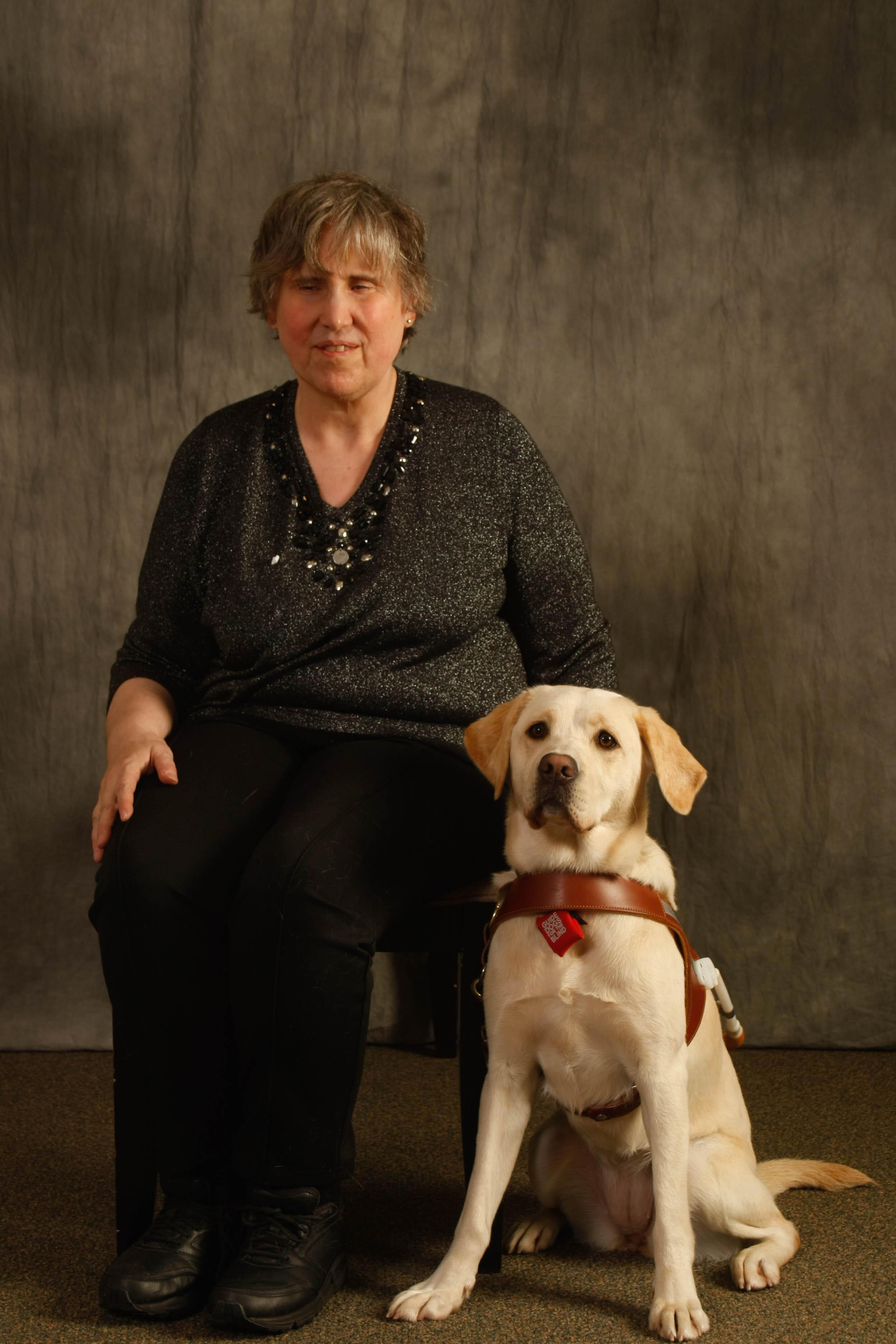 Class photo of Deb seated beside yellow Lab guide dogs Praline at their GDB graduation.
