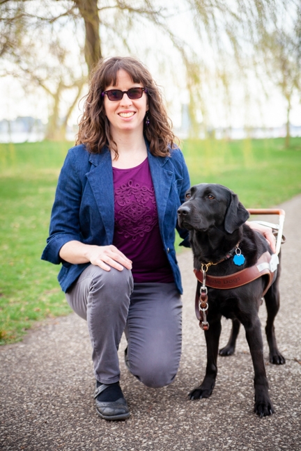 A portrait of Leanne Bremner kneeling next to her black Lab guide dog, Eva.