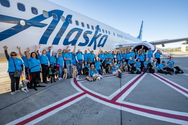 A group photo of people and puppies on the tarmac in front of an Alaska Airlines plane.
