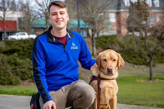Guide Dog Mobility Instructor Joey Foat kneels next to a yellow Lab guide dog.