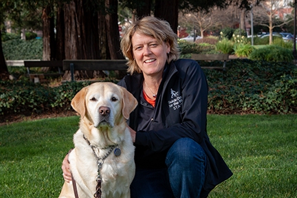 Dr. Kate Kuzminski kneeling next to a yellow Lab on a green grass with trees in the background.