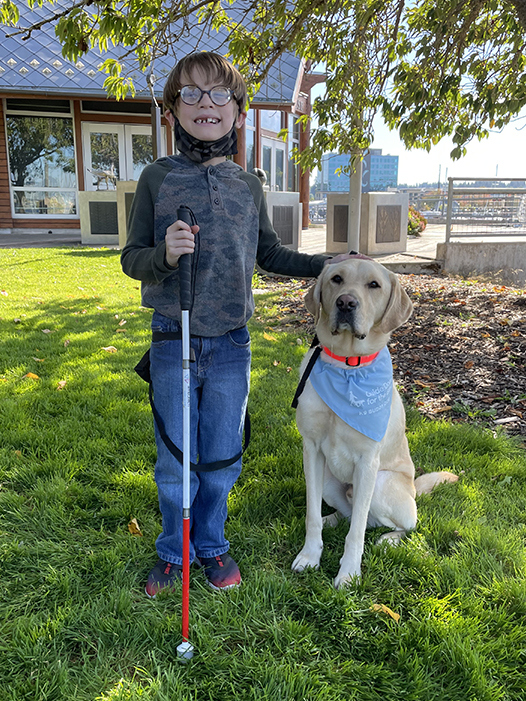 A young boy with a white cane stands next to a yellow Lab K9 Buddy dog.