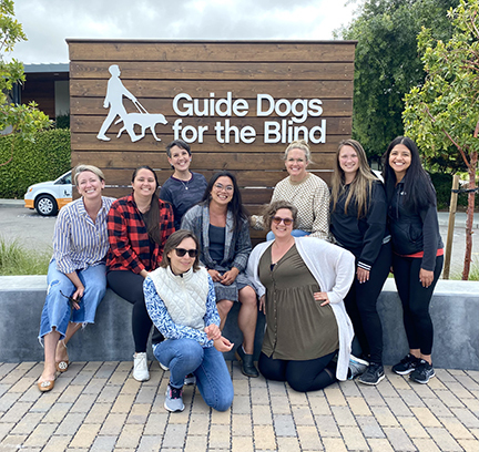 A group of nine university students in front of a Guide Dogs for the Blind logo sign.