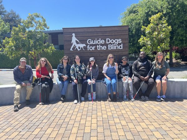 Workshop attendees are seated together in front of the GDB sign on a sunny day. Some attendees hold their white canes in front of them. All are smiling at the camera.