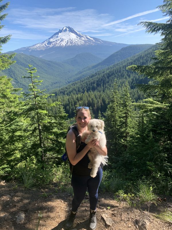 Kadi smiles for a photo op on a hiking trail with stunning mountain vistas. Kadi is holding a small white dog in her arms.