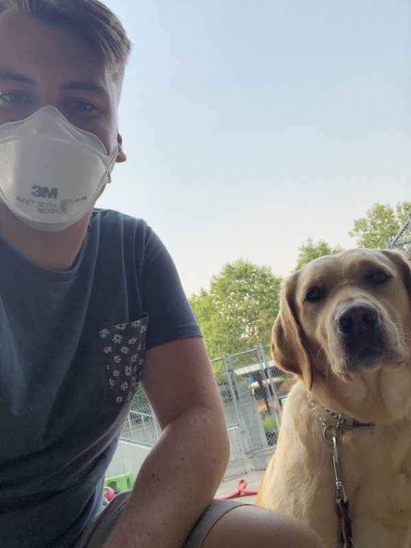 GDB Trainer Joey Float kneels beside a yellow Lab guide dog with smokey skies behind them. Joey wears an N95 mask to protect himself from smoke inhalation.