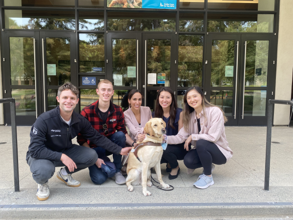 Five Ophthalmology residents kneel together on the GDB campus with a yellow Lab guide dog in harness.