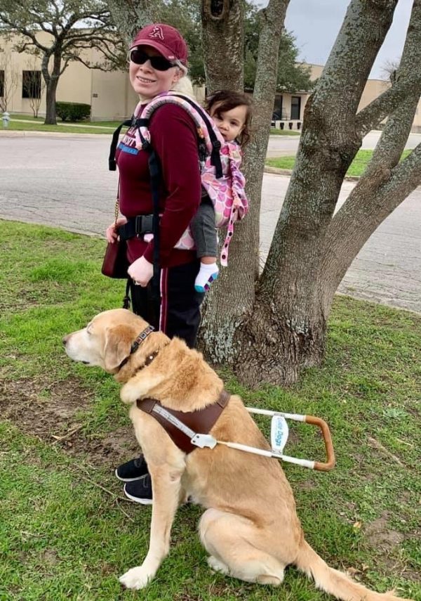 Melissa Padron with her guide dog Cameo. Melissa carries her young daughter in a baby carrier on her back.