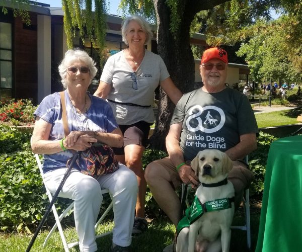 Three puppy raising volunteers post next to a yellow Lab guide dog puppy at Fun Day.