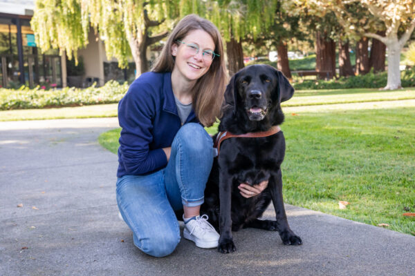 Dr. Sarah Elizabeth Byosiere kneels next to a black Lab guide dog.