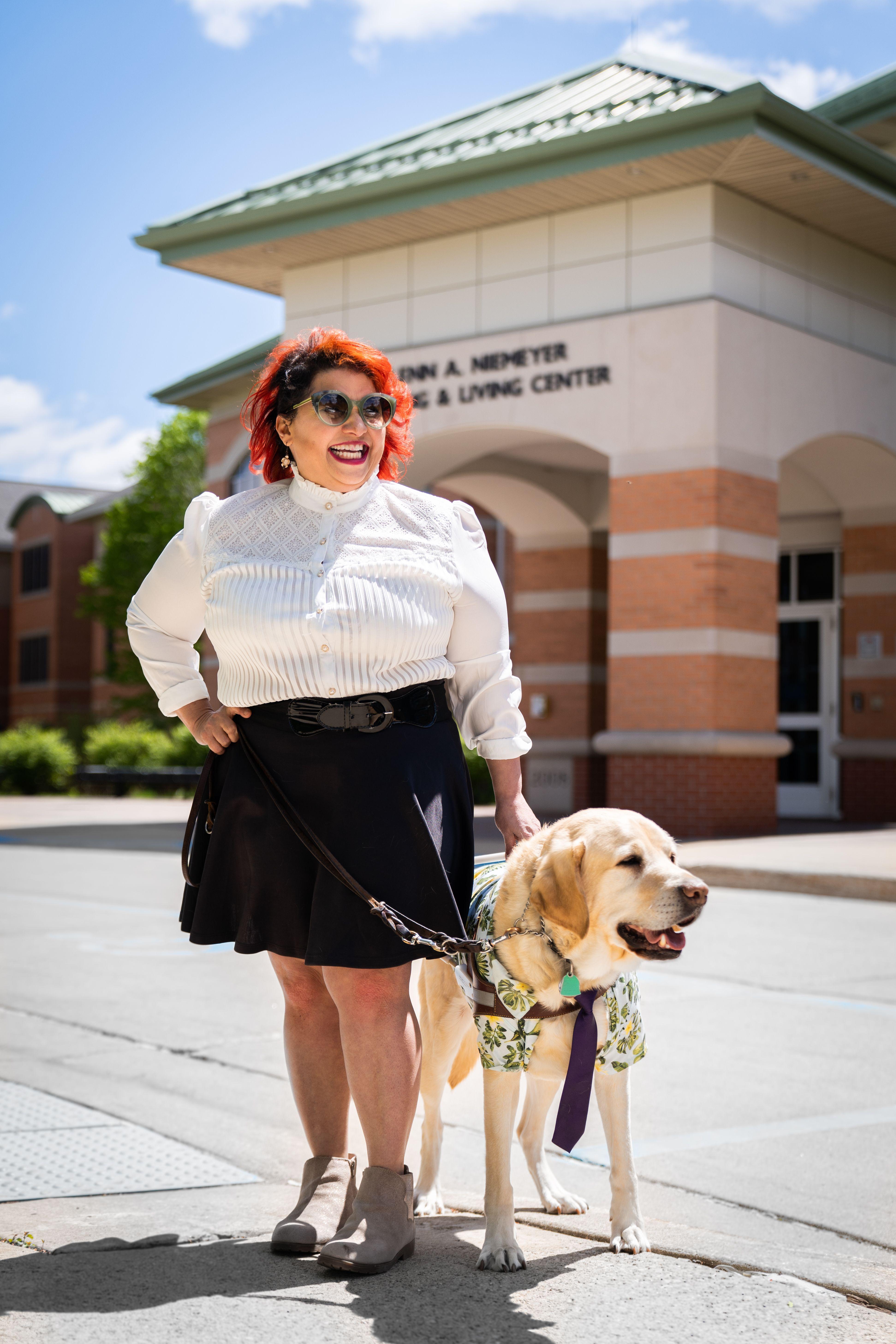 Melba stands with her yellow Lab guide dog Chad on the campus of the college where she is tenured professor. She  smiles widely at the camera.