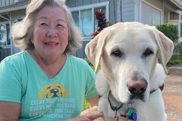 A woman sits beside her yellow Lab guide dog in front of their house.