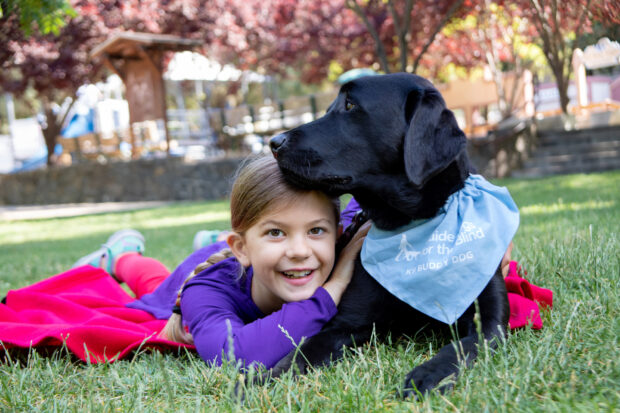 A young girl lays next to her black Lab K9 Buddy dog on the grass at a park.