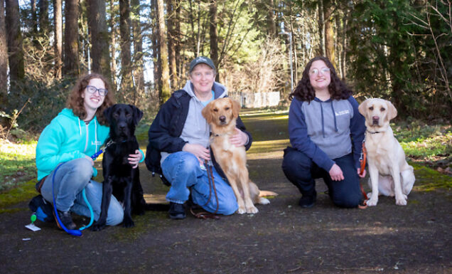 Three kennel techs kneel on a tree-lined walking path with three dogs in training.