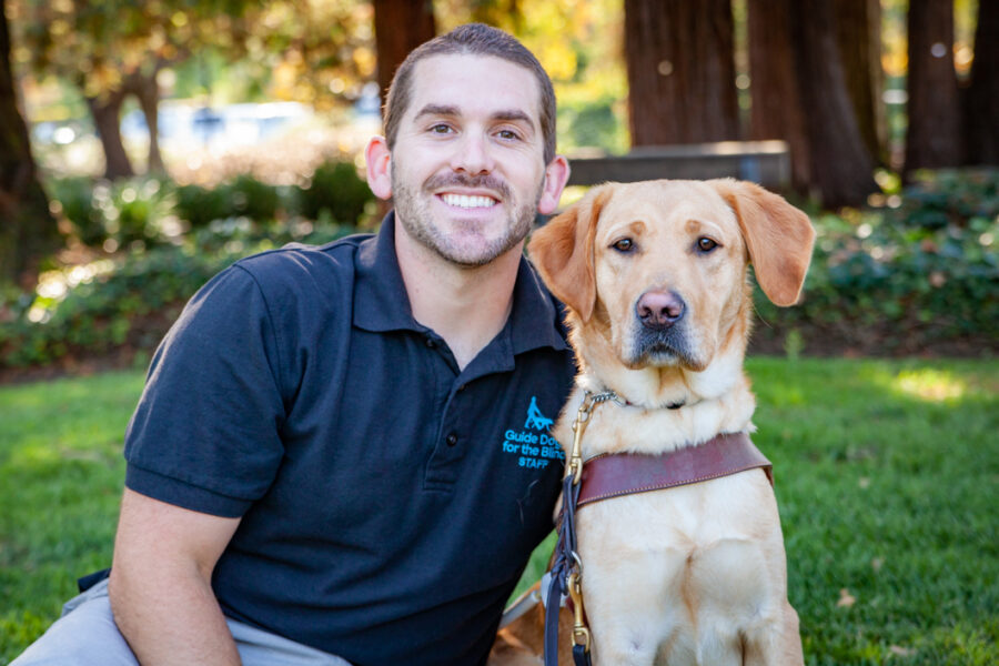 A man sits on a green lawn next to a yellow Lab guide dog.