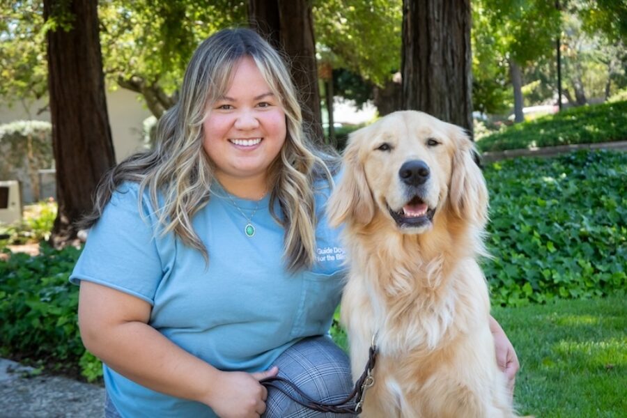 A woman kneels beside a smiling Golden Retriever.