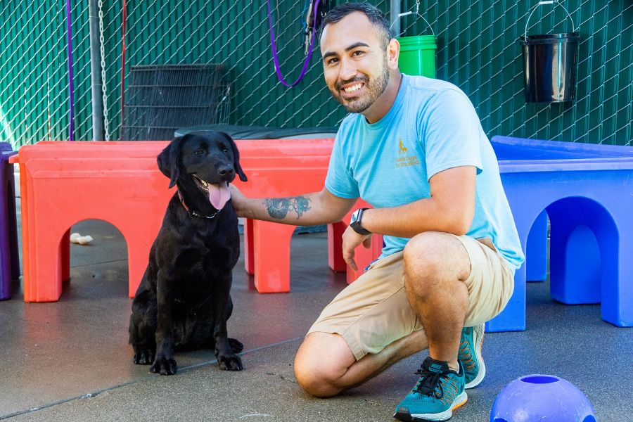 A portrait of Bobby with a black Lab in a dog play area full of colorful toys.