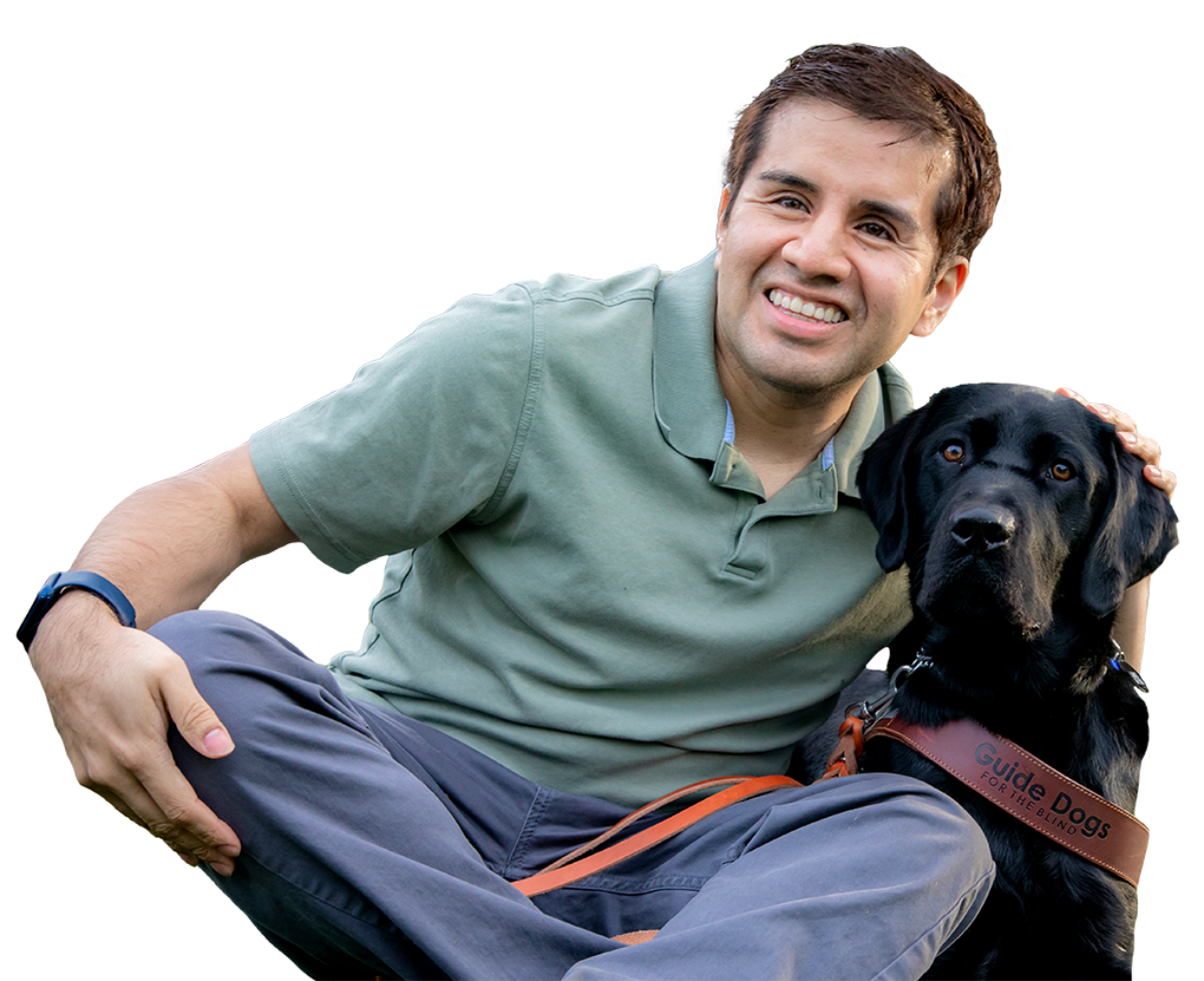 A smiling man sits next to and embraces his black Lab guide dog.