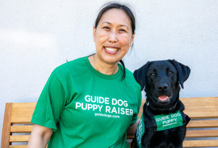A woman wears a green shirt that says "Guide Dog Puppy Raiser" and sits on a bench next to a black Lab guide dog puppy in training wearing their green vest.