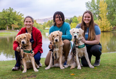 Three Guide Dogs for the Blind volunteer puppy raisers pose with three Golden Retriever guide dog puppies lakeside in a park.