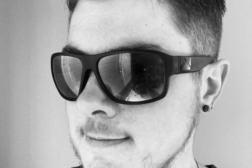 Black and white image of Ness. He is wearing a black tee and sunglasses with a slight smile on his face.