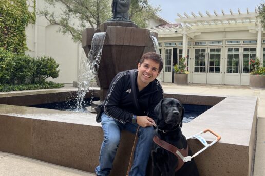Belo is seated beside a fountain with his black Lab guide dog, Limo by his side.