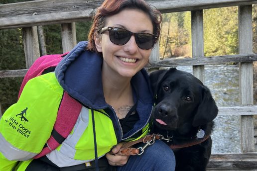 Brooke kneels beside her black Lab guide dog on a bridge. Brook is wearing her reflective GDB jacket and a red backpack. The dog looks curiously at the camera.