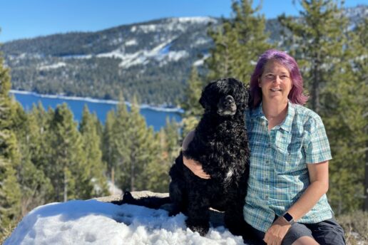 Jenna with her black pet dog in the snow above a blue lake.