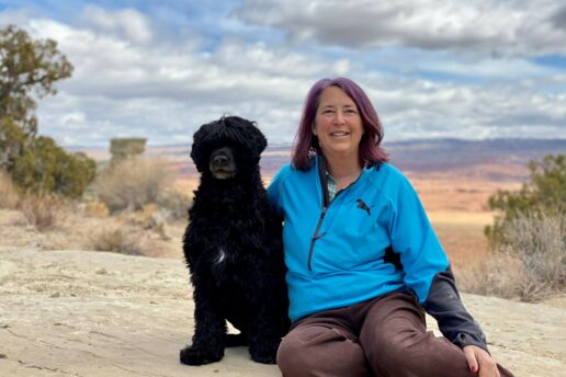Jenna is seated beside her pet dog in front of a high desert vista.