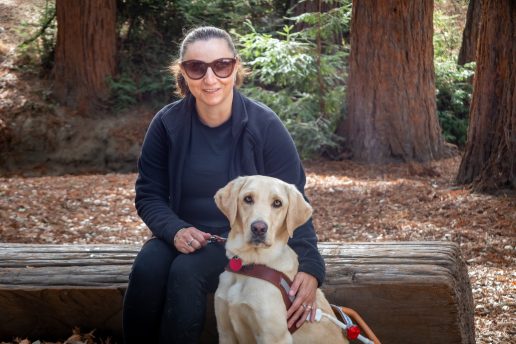 Kate sits on a log beside her yellow Lab guide dog. She rests one hand on the dog's shoulder and holds the leash with the other.
