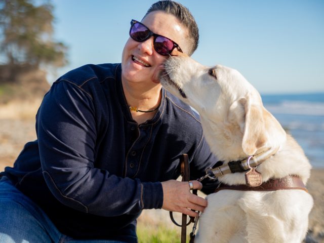 Cheri is seated beside her yellow Lab guide dog, Martinez, near the coast. Martinez lands a big kiss on her cheek while Cheri laughs.