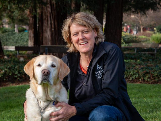 Dr. Kate kneels beside a yellow Lab. Kate is wearing a black Staff jacket and the dog looks seriously at the camera.