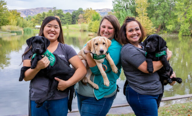 Three women stand in front of a lake in a park and each person holds a young guide dog puppies in her arms.