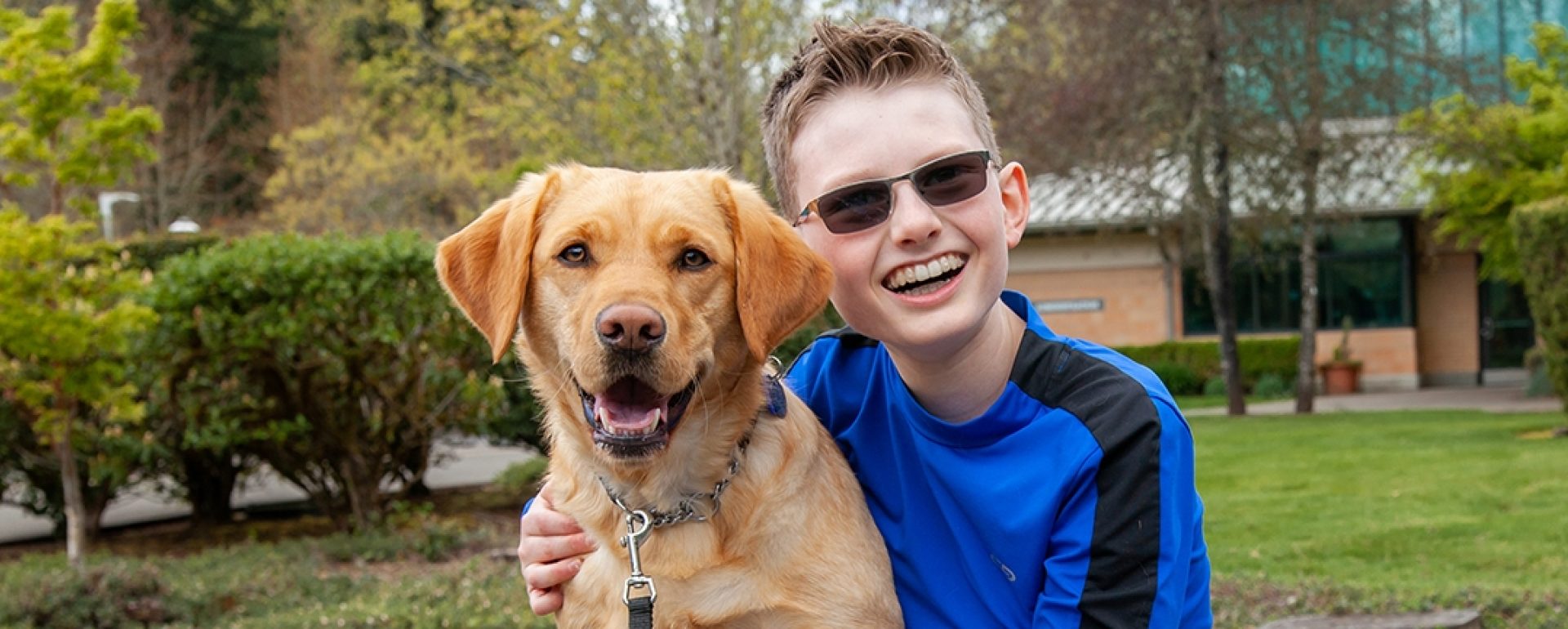 A young boy hugging a yellow Lab K9 Buddy dog; both have ear-to-ear grins.
