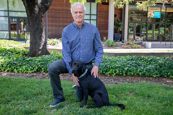 Tom Horton kneels in the grass next to a black Lab