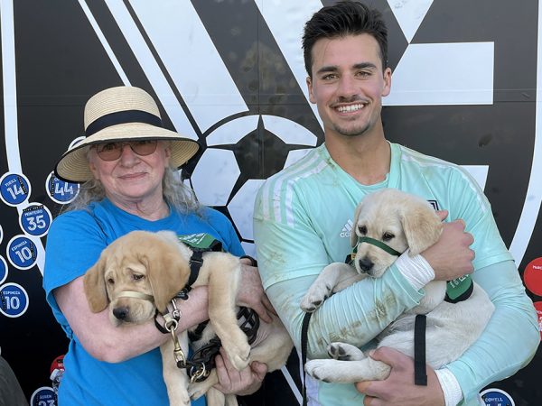 A GDB puppy raising volunteer and San Jose Earthquakes Goalie JT Marcinkowsi both hold yellow Lab guide dog puppies.